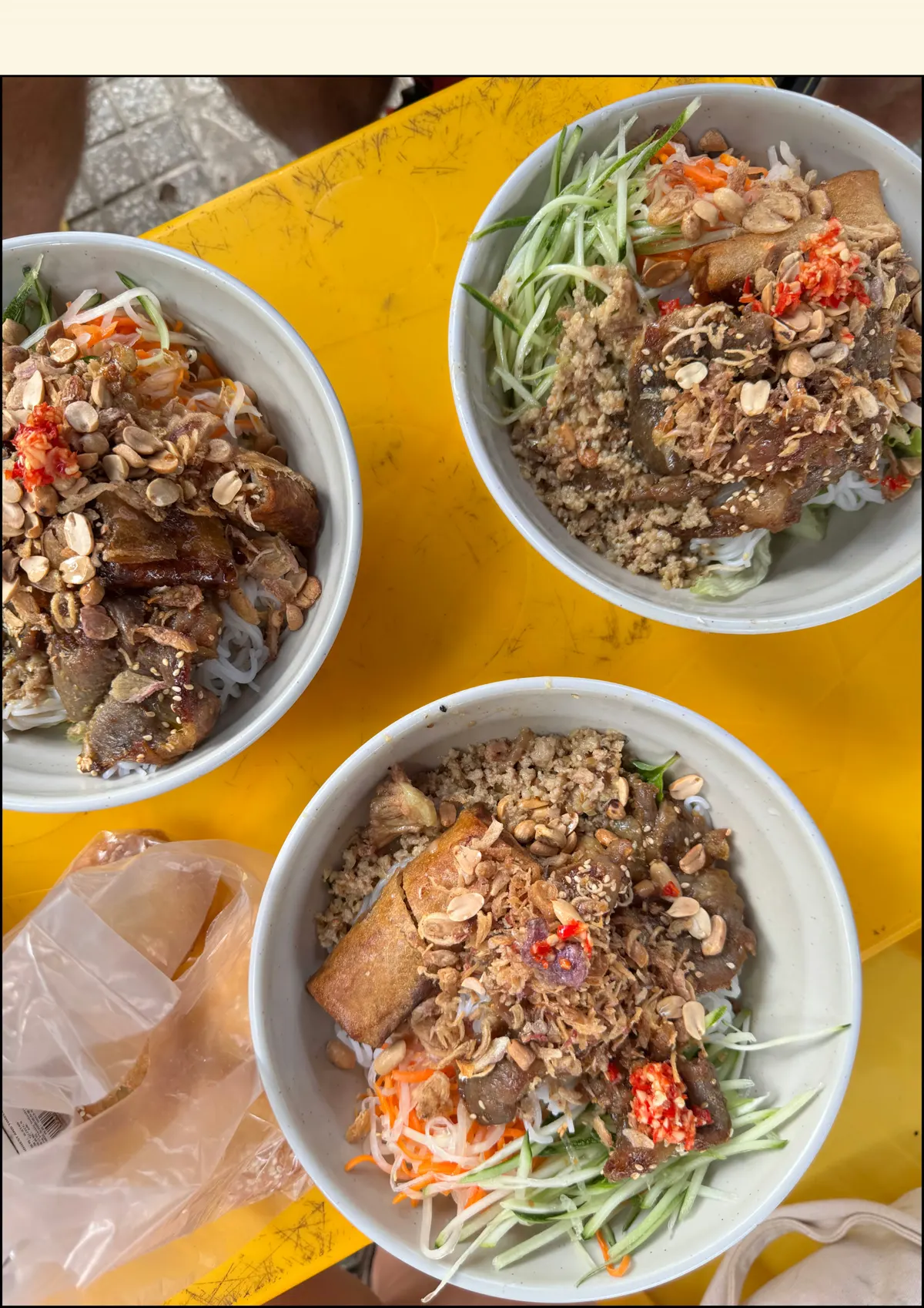 Three bowls of noodles topped with herbs, peanuts and spring rolls on a yellow table, seen from above