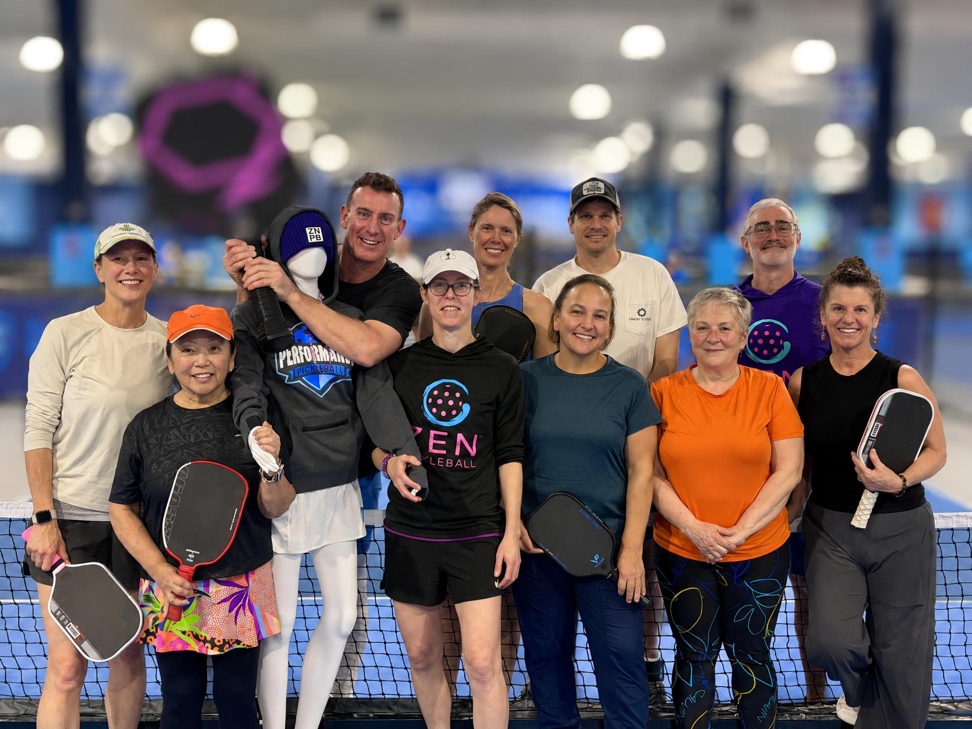 Rachel Rhoney standing behind a court net with a group of her pickleball clients, most holding paddles and smiling at the camera