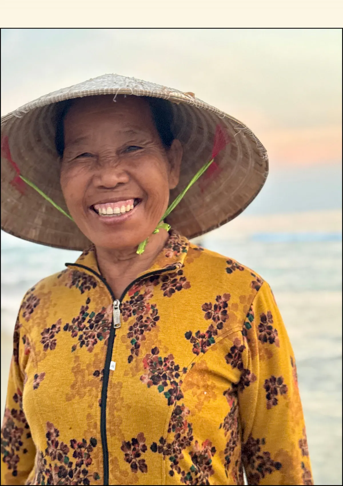 A smiling Vietnamese woman in a conical hat and a floral jacket at dusk